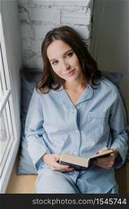Beautiful relaxed woman with carefree expression, dressed in nightclothes, reads book on window sill in morning, enjoys spare time, likes her hobby. Vertical shot of teenage girl with literature