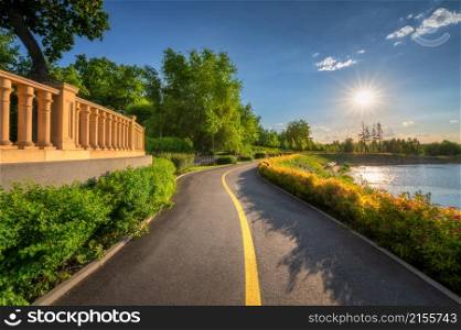 Beautiful road in park at sunset in summer. Colorful landscape with walkway, green bushes, flowers, trees, lake and blue sky with sun at sunny bright day. Bike road in blooming park. Walk. Nature