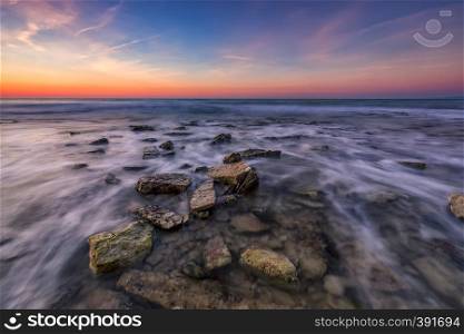 Beautiful rocky beach with stones in blurred water, the colorful sky at sunrise. Black sea, Bulgaria. Landscape with the sea waves and rocks.