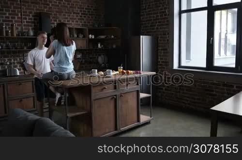 Beautiful romantic couple hugging while spending time together in modern kitchen in the morning. Back view of female sitting on kitchen table while handsome man embracing her and gently kissing. Slow motion. Steadicam stabilized shot.