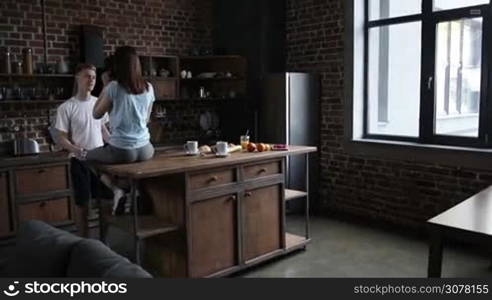 Beautiful romantic couple hugging while spending time together in modern kitchen in the morning. Back view of female sitting on kitchen table while handsome man embracing her and gently kissing. Slow motion. Steadicam stabilized shot.