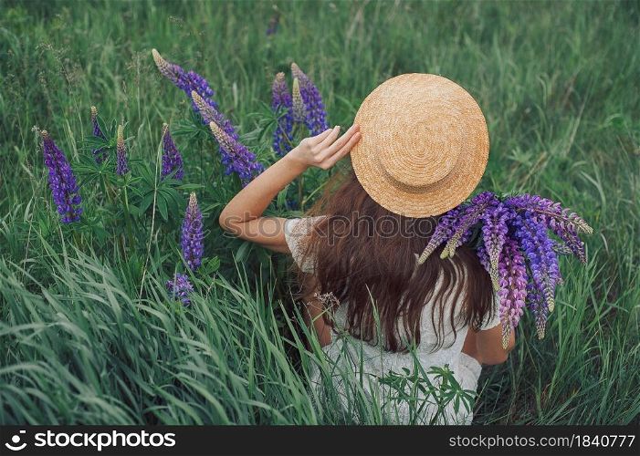 Beautiful romantic woman with bouquet of lupines joyfully in white dress and hat sits in field of purple lupine flowers. Soft selective focus.. Beautiful romantic woman with bouquet of lupines joyfully in white dress and hat sits in field of purple lupine flowers. Soft selective focus
