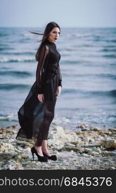 Beautiful sad gothic woman in black dress standing on the sea beach