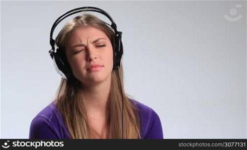 Beautiful sad pensive young woman in headphones listening music on white. Attractive brunette girl moving slowly to music being gloomy and thoughtful, looking at camera with eyes full of sadness, closing her eyes and putting hand on headphones.