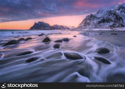 Beautiful sandy beach with stones in blurred water, colorful cloudy pink sky and snowy mountains at sunset. Utakleiv beach, Lofoten islands, Norway. Winter landscape with sea, waves, rocks at dusk