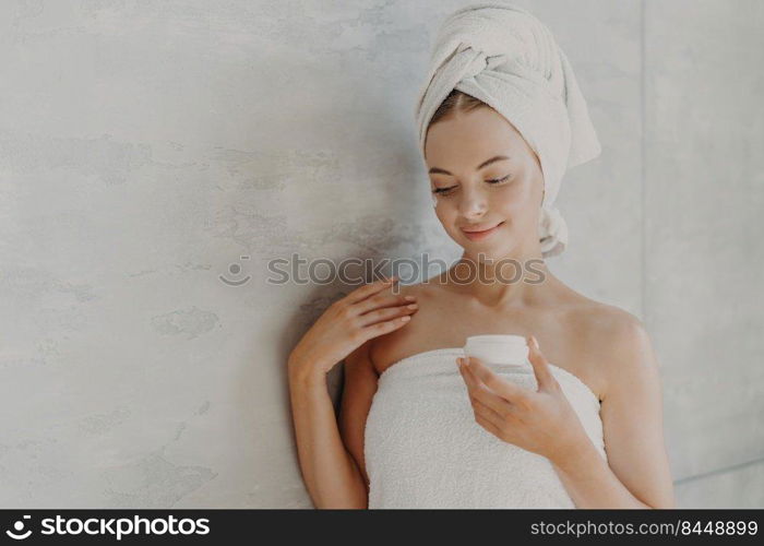 Beautiful satisfied woman with healthy skin, minimal makeup, applies face cream, stands wrapped in bath towel near grey background after taking shower. Sauna morning routine, skin care concept