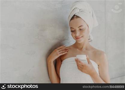 Beautiful satisfied woman with healthy skin, minimal makeup, applies face cream, stands wrapped in bath towel near grey background after taking shower. Sauna morning routine, skin care concept