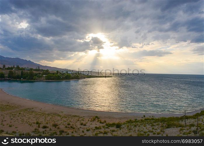 Beautiful scenic view - distant open wood amid the calm water of Issyk-Kul Lake against the background of Tien Shan mountain range and cloudy blue sky, Kyrgyzstan, Central Asia