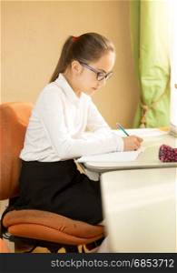 Beautiful schoolgirl wearing eyeglasses posing behind desk while doing homework