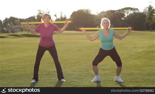 Beautiful senior blonde females with bodybars on shoulders doing squat exercises in park in rays of setting sun. Charming senior fit women in sportswear squatting with body bar, making exercises for hips and buttocks while training outdoors.