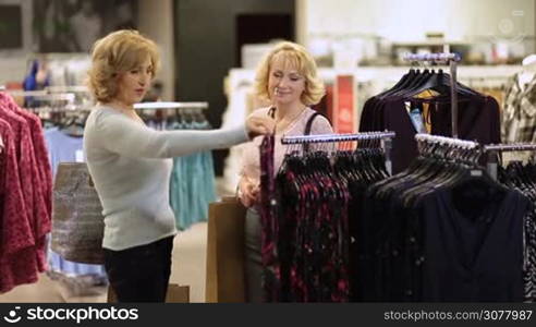 Beautiful senior blonde selecting apparel with her girlfriend while shopping for clothes in fashion store. Two attractive elderly women with shopping bags looking at each other with smile while standing near hangers at clothing store.