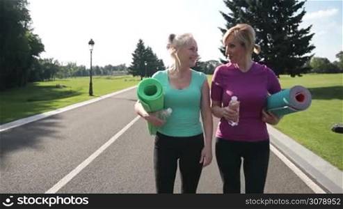 Beautiful senior females in sport clothes holding exercise mats going through the park to make sport. Slim fitness adult women preparing to workout outdoors, walking in the park and talking. Slow motion. Stabilized shot.