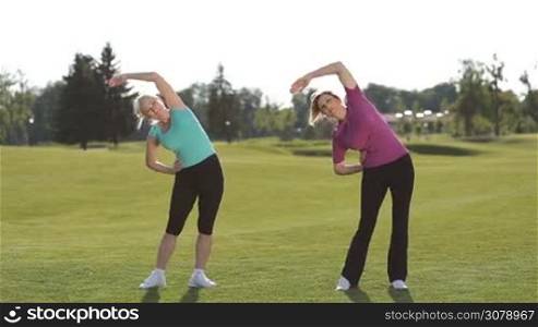 Beautiful senior fitness women in sportswear doing stretching exercises and warmup while standing on park lawn. Attractive active adult blonde females stretching arms and shoulders while working out outdoors in park on a sunny day.