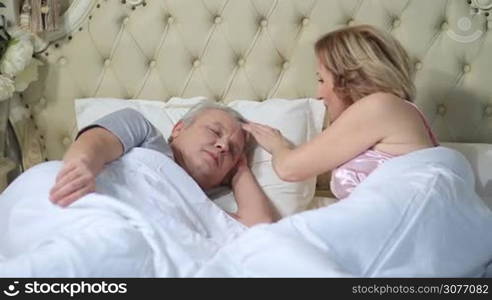 Beautiful senior woman waking up her husband in the morning tenderly caressing his head. Mature man waking up and stretching in the bed while his wife touching him gently with love.