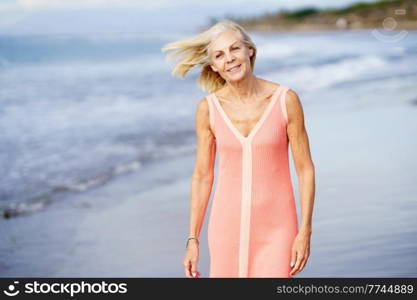 Beautiful senior woman walking along the shore of a tropical beach, wearing a nice orange dress. Elderly female enjoying her retirement at a seaside retreat.. Elderly female enjoying her retirement at a seaside retreat. Senior woman on shore of a beach.