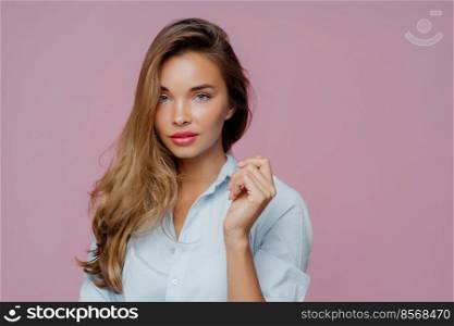 Beautiful serious European woman keeps arm raised, looks confidently at camera, has well cared face, makeup, dressed in shirt, has natural beauty, models against purple background copy space for promo