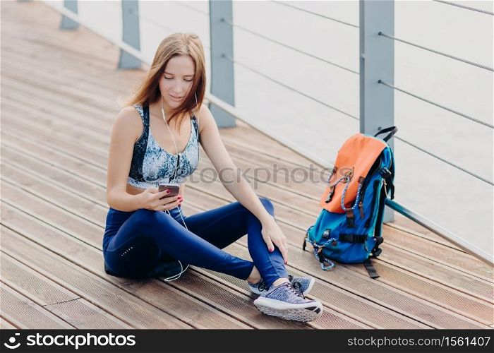 Beautiful serious female has gaze at screen of smart phone, checks which song to listen, uses modern earphones for entertainment, sits crossed legs near bag outdoor, has rest after training.