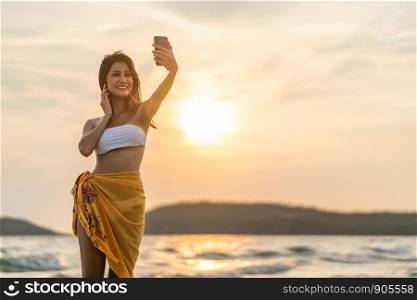 Beautiful sexy Asian girl smiling taking selfie photo or live video call, using smartphone on beach vacation at sunset. Asia travel, holiday trip, leisure activity or happy people lifestyle concept