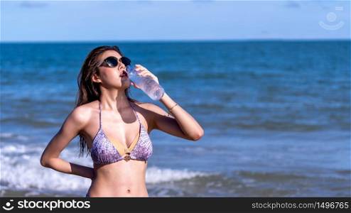 beautiful, sexy Asian woman wearing swimsuit and sunglasses, drinking cold mineral water on the beach in the summer on the weekends.