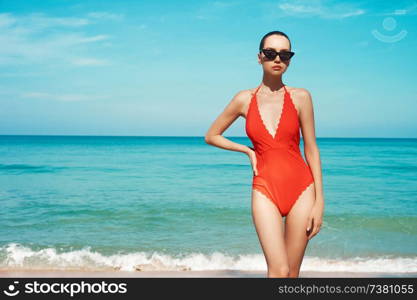 Beautiful sexy lady on tropical beach. Fashionable woman with slim perfect figure walking in front of blue sea. Model pose in red swimwear and modern sunglasses. Relax portrait of young sunbathing beauty