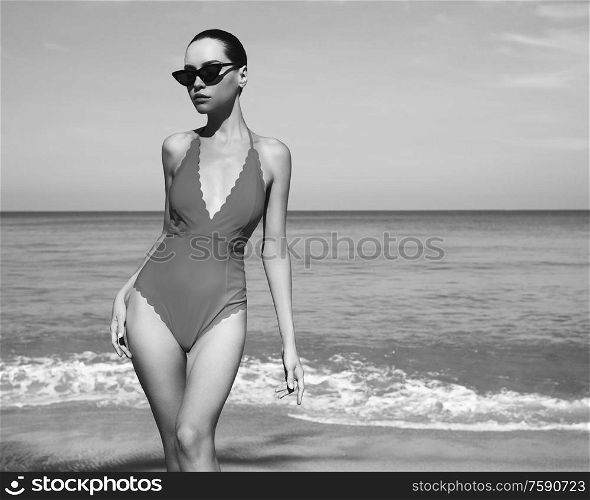 Beautiful sexy lady on tropical beach. Fashionable woman with slim perfect figure walking in front of blue sea. Model pose in swimwear and modern sunglasses. Relax portrait of young sunbathing beauty