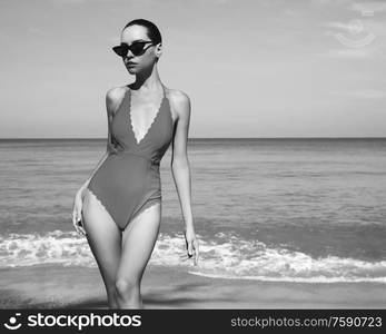 Beautiful sexy lady on tropical beach. Fashionable woman with slim perfect figure walking in front of blue sea. Model pose in swimwear and modern sunglasses. Relax portrait of young sunbathing beauty