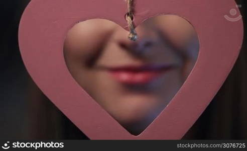 Beautiful sexy pink lips giving kiss through the heart-shaped frame. Woman&acute;s perfect lips giving air kiss. Shallow depth of field with focus on pink heartshape frame hanging in front of camera.