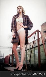 Beautiful sexy young girl in fur coat and lingerie stands at railroad station. Full length
