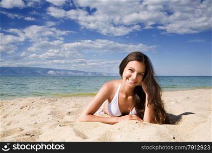Beautiful sexy young woman with long brunette hair lying down at the beach in the sunny day and looking at camera