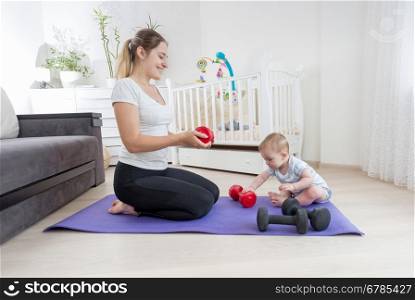 Beautiful slim mother with her baby son sitting on fitness mat at home and exercising with dumbbells