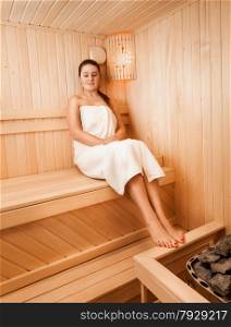 Beautiful slim woman in towel sitting on bench at sauna next to oven