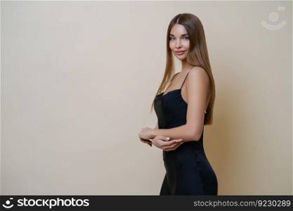 Beautiful slim woman stands in profile, wears elegant black dress, has long straight hair, looks with charming expression at camera, poses against beige background, copy space for your information