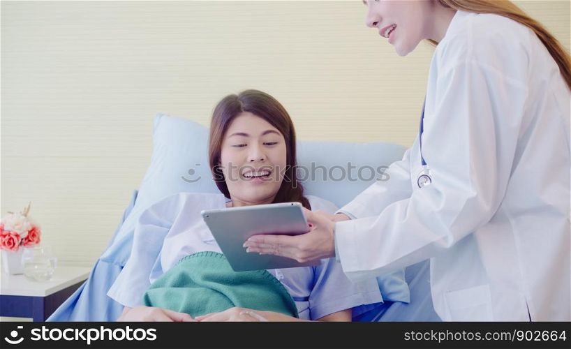 Beautiful smart Asian doctor and patient discussing and explaining something with tablet in doctor hands while staying on Patient's bed at hospital. Medicine and health care concept.