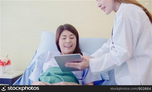 Beautiful smart Asian doctor and patient discussing and explaining something with tablet in doctor hands while staying on Patient's bed at hospital. Medicine and health care concept.