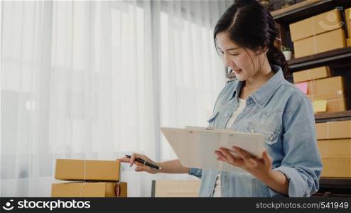 Beautiful smart Asian young entrepreneur business woman owner of SME checking product on stock and write on clipboard working at home. Small business owner at home office concept.