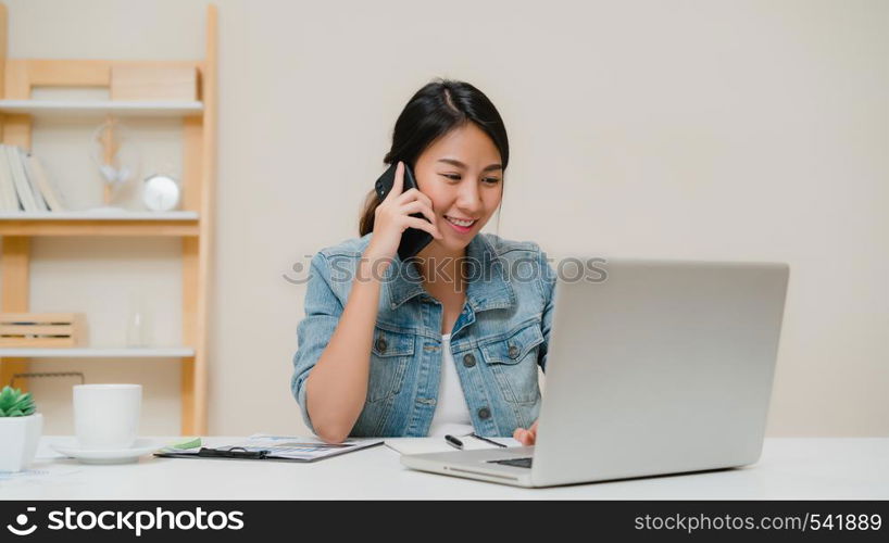 Beautiful smart business Asian woman in smart casual wear working on laptop and talking on phone while sitting on table in creative office. Lifestyle women working at home concept.