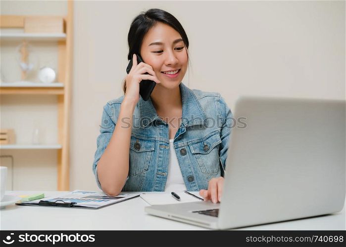Beautiful smart business Asian woman in smart casual wear working on laptop and talking on phone while sitting on table in creative office. Lifestyle women working at home concept.