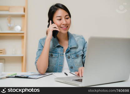 Beautiful smart business Asian woman in smart casual wear working on laptop and talking on phone while sitting on table in creative office. Lifestyle women working at home concept.