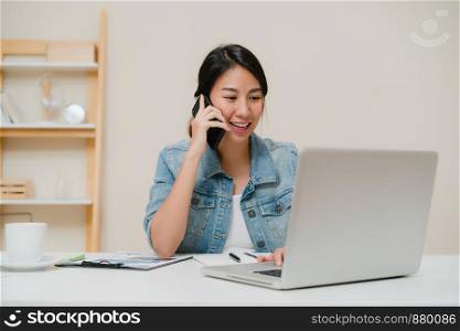 Beautiful smart business Asian woman in smart casual wear working on laptop and talking on phone while sitting on table in creative office. Lifestyle women working at home concept.