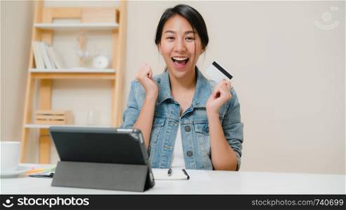 Beautiful smart business Asian woman using tablet buying online shopping by credit card while wear smart casual sitting on desk in living room at home. Lifestyle woman working at home concept.