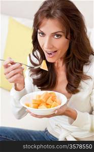Beautiful smiling brunette woman at home sitting on sofa or settee eating a bowl of fresh cut melon fruit.