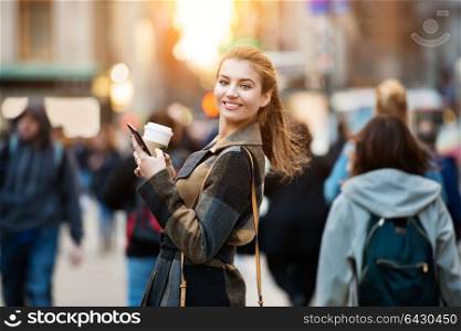 Beautiful smiling businesswoman walking on busy city street from work with coffee cup and texting on mobile phone at sunset time.