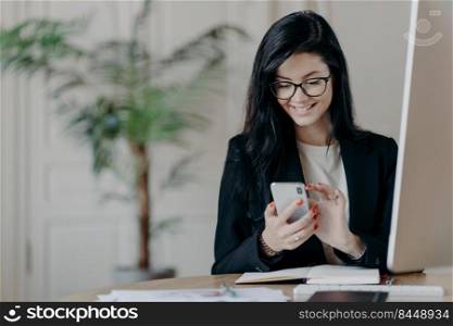 Beautiful smiling female freelancer works remotely, concentrated in smartphone, sends feedback on received message, sits in coworking space, dressed formally, works on computer, surfs internet