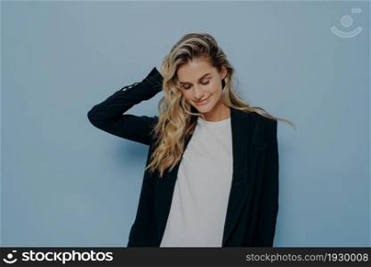 Beautiful smiling female with blonde dyed hair feeling happy, wearing black blazer over white tshirt while standing isolated against blue background, positive woman keeping eyes closed and smiling. Beautiful smiling female with blonde dyed hair feeling happy while posing in studio