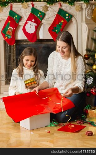 Beautiful smiling girl and mother wrapping Christmas gifts in red paper and tying with golden ribbon