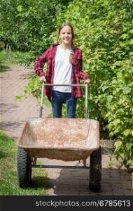 Beautiful smiling girl in jeans and shirt with wheelbarrow at rustic garden