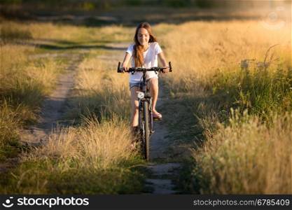 Beautiful smiling girl riding on bicycle in meadows at sunset