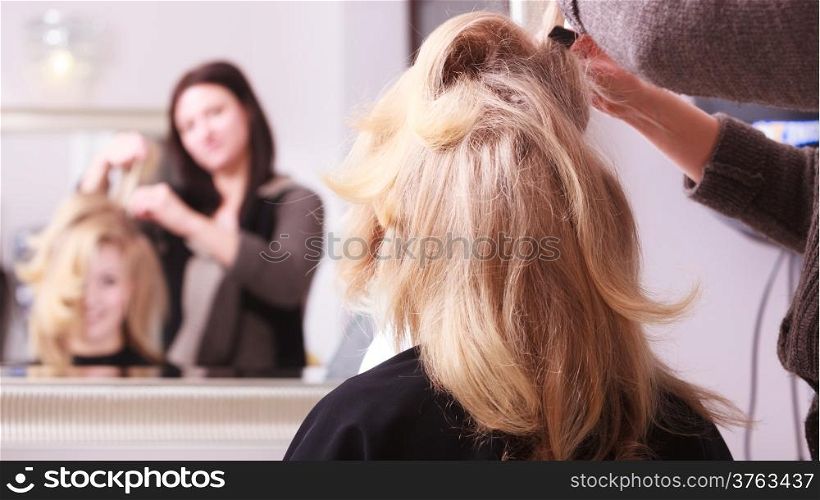 Beautiful smiling girl with blond wavy hair by hairdresser. Hairstylist combing female client young woman in hairdressing beauty salon. Hairstyle.