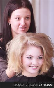 Beautiful smiling girl with blond wavy hair by hairdresser. Hairstylist combing female client young woman in hairdressing beauty salon. Hairstyle.