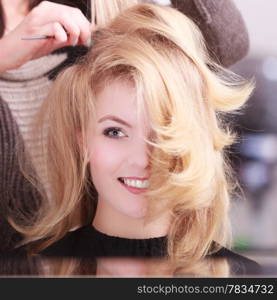 Beautiful smiling girl with blond wavy hair by hairdresser. Hairstylist with comb combing female client young woman in hairdressing beauty salon. Hairstyle.